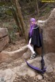 A woman with purple hair standing on a rock in the woods.